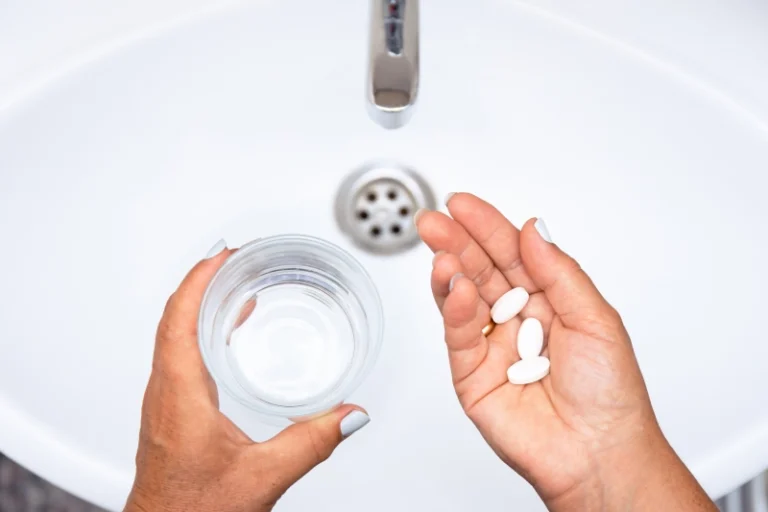 Una persona sostiene unas pastillas en una mano mientras que en la otra tiene un base de agua sobre le labavo.