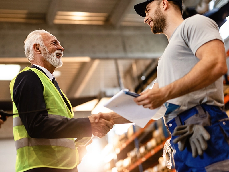 Un hombre mayor con barba y chaleco de alta visibilidad se da la mano con un trabajador de una almacén de material.